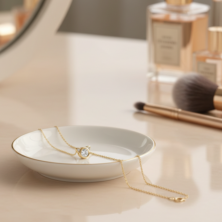 A delicate gold necklace with a small oval gemstone pendant is draped over a white ceramic dish with a gold rim, set on a light beige tabletop with blurred cosmetic items in the background.